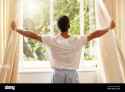 back-view-man-and-open-curtain-at-window-to-good-morning-sunrise-and-sunshine-to-relax-in-room-at-home-guy-wake-up-and-opening-bedroom-drapes-in-2RD55KN