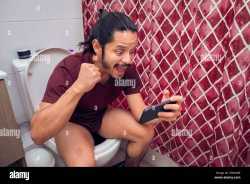 young-latin-man-smiling-on-the-toilet-using-a-smart-phone-at-the-bathroom-2F8X4HR